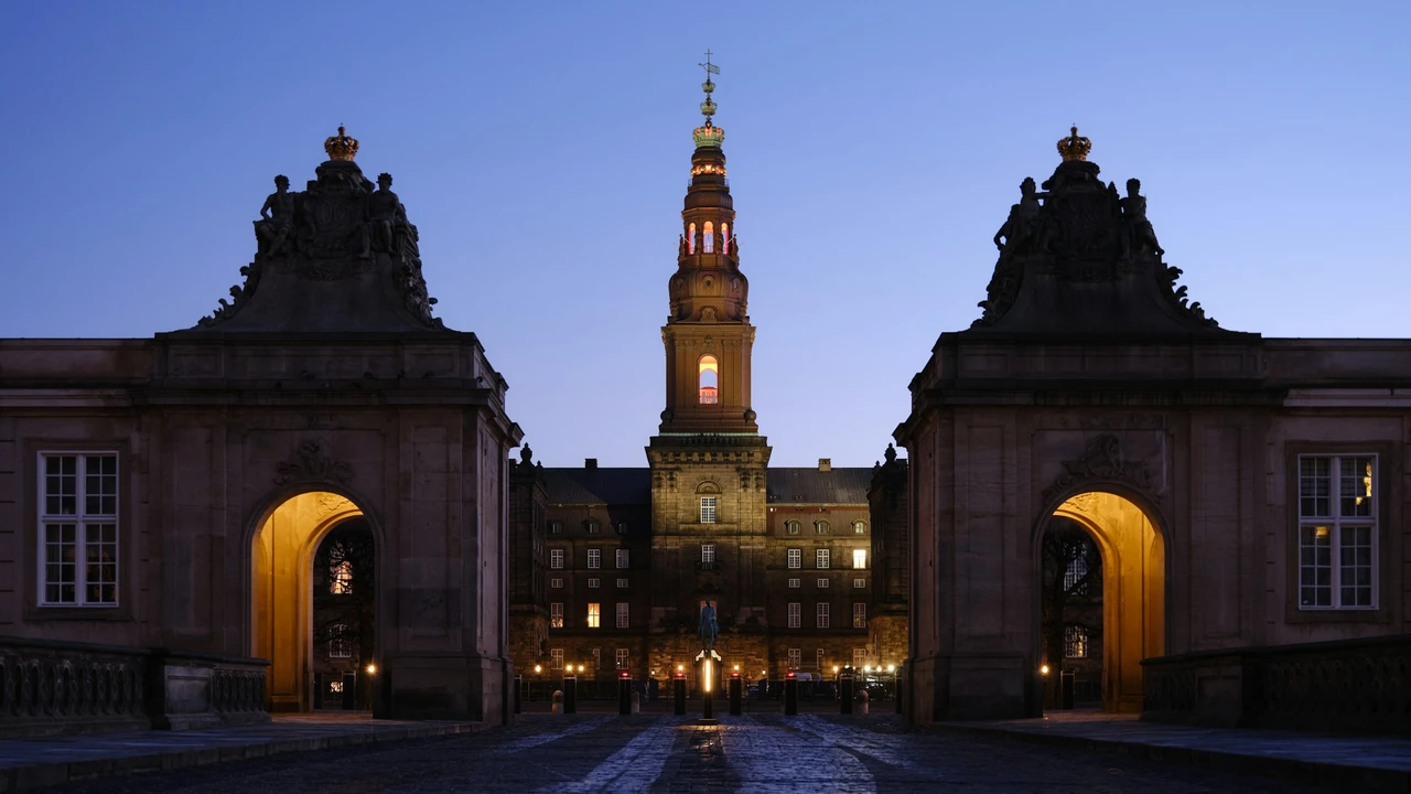 A symmetrical, low-angle shot of Christiansborg Palace in Copenhagen at dusk, framed by two large stone gatehouses with lit archways in the foreground. The central tower of the palace is illuminated, glowing against a deep blue evening sky, with its ornate green and gold spire reaching upward.