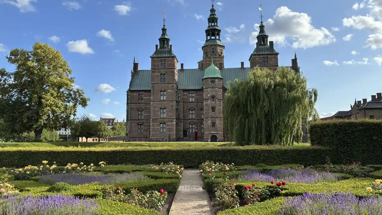 A medium-long shot of Rosenborg Castle in Copenhagen, Denmark, a Dutch Renaissance-style palace featuring red brick, slim towers with green copper spires, and a central tiered steeple. The castle is set behind a manicured Royal Garden in the foreground, showcasing neat rows of purple lavender and red roses separated by a gravel path.