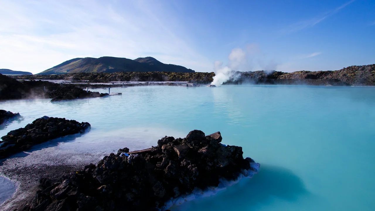 A wide landscape shot of the Blue Lagoon in Iceland, featuring milky-blue geothermal water surrounded by dark, rugged volcanic lava rocks under a clear sky, with steam rising from the water's surface in the distance