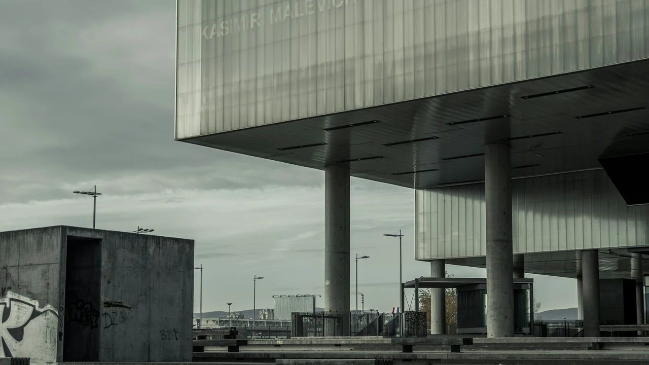 A wide, low-angle shot of the modern Museum of Contemporary Art (MSU) in Zagreb, Croatia, under a moody, overcast sky. The building features a large, cantilevered upper floor with a translucent glass facade supported by thick concrete pillars. The name 