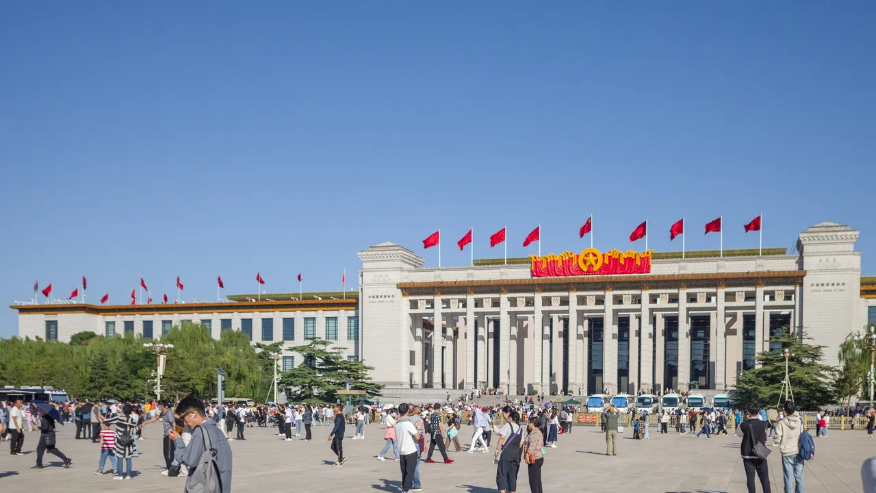 A wide-angle, eye-level shot of the National Museum of China in Beijing. The massive, neoclassical building features a long facade with light-colored stone columns and is topped with several bright red flags fluttering against a clear blue sky. In the foreground, a large, open stone plaza is bustling with many tourists walking about. A prominent red and gold emblem is displayed at the center of the building's roofline.