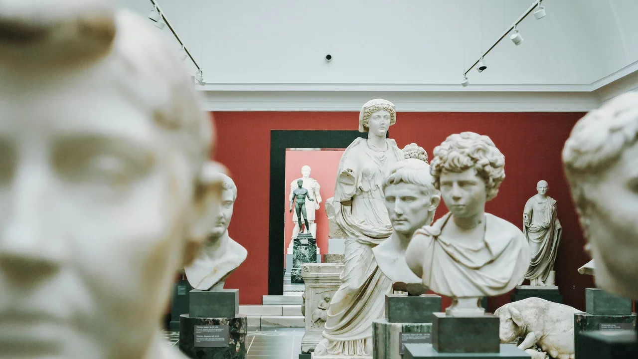 An eye-level, medium shot inside an exhibit at the National Museum of Denmark, showcasing a collection of ancient Roman marble sculptures. Several white marble portrait busts are mounted on dark pedestals in the foreground and middle ground, some blurred to create depth. In the center, a full-body female statue draped in robes stands before a deep red wall, while more statues are visible through a doorway in the background under a bright, skylit ceiling.