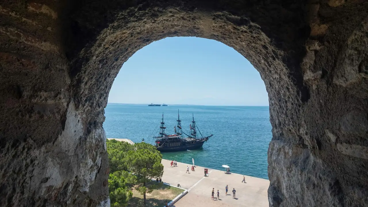 View through the Arch of Galerius in Thessaloniki, overlooking the seaside promenade and the Aegean Sea with a pirate-style tour boat anchored offshore.