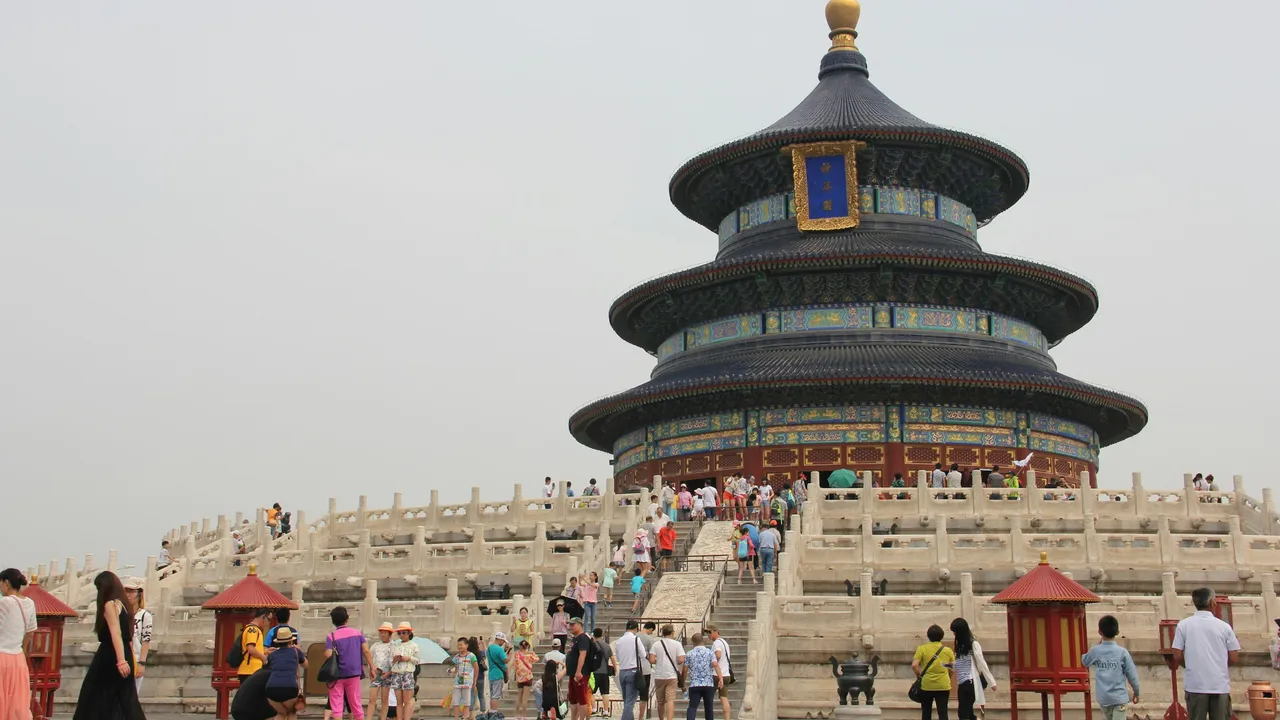 A wide-angle shot of the Temple of Heaven in Beijing, featuring the circular, blue-roofed Hall of Prayer for Good Harvests standing atop a three-tiered white marble terrace, with numerous tourists walking around the base on a cloudy day.