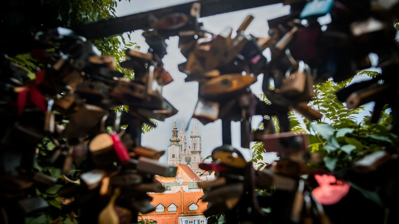 A creative, framed shot of the Zagreb Cathedral in Croatia seen through a gap in a metal fence covered in out-of-focus love locks. The iconic twin spires and the white tower of the cathedral are centered in the distance under a pale sky. In the mid-ground, traditional orange-tiled roofs and green trees are visible, while the foreground is dominated by the blurred silhouettes of numerous padlocks attached to the railing.
