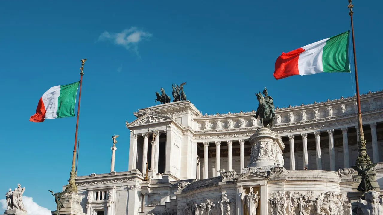 Italian national flags flying in front of the Altare della Patria (Vittoriano Monument) in Rome, Italy, featuring classical white marble architecture, statues, and a clear blue sky.