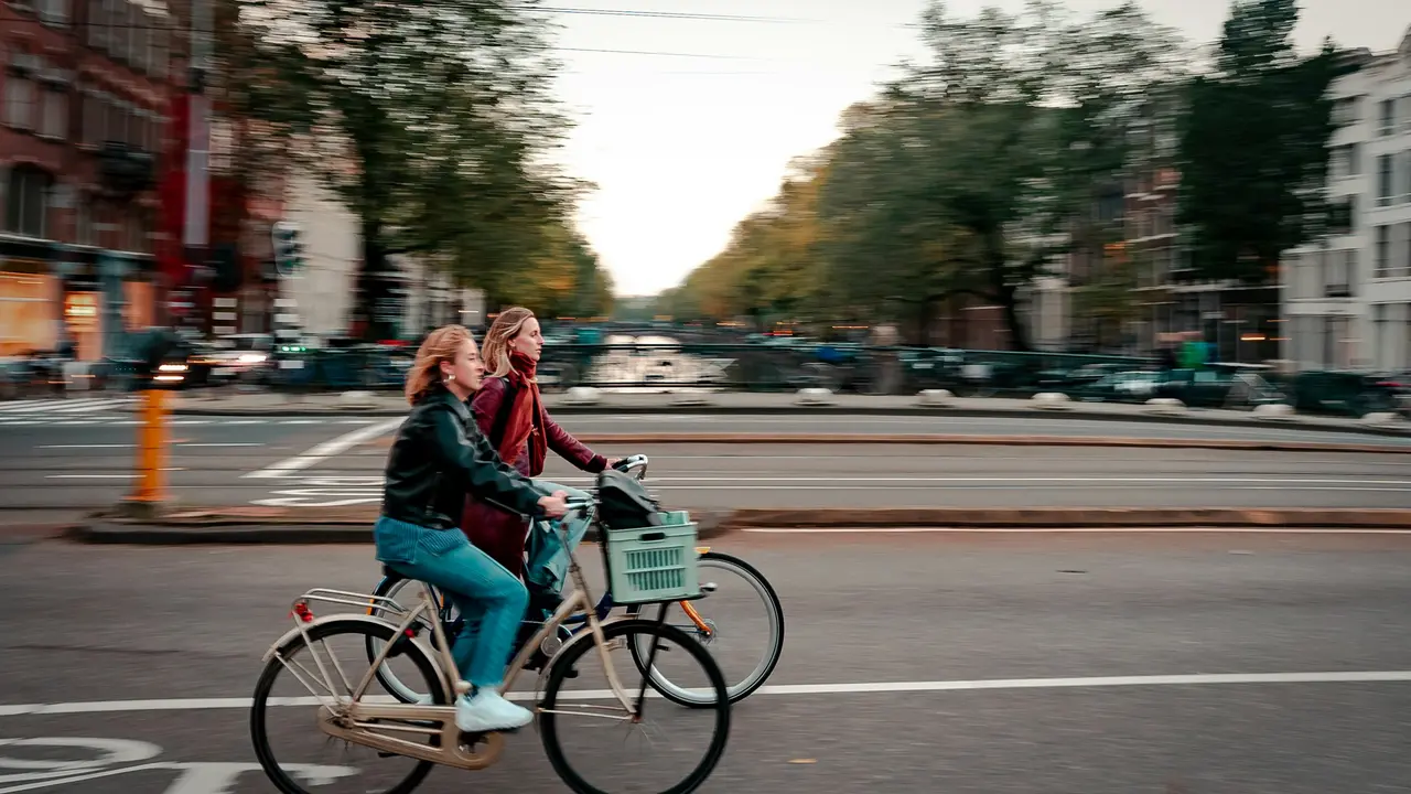Two women riding bicycles along a tree-lined city canal at sunset with urban buildings and street traffic in the background