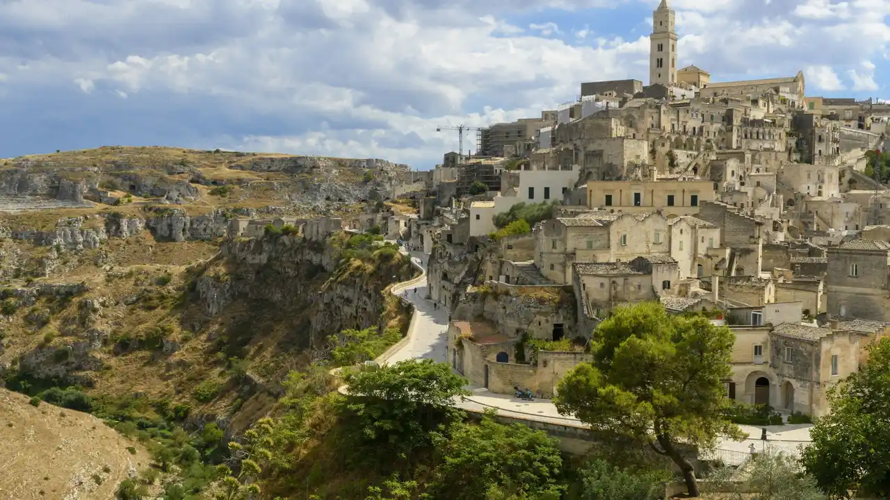 Panoramic view of Matera, Italy, showcasing the ancient Sassi cave dwellings carved into limestone cliffs, historic stone houses, winding roads, and a church tower overlooking the ravine.