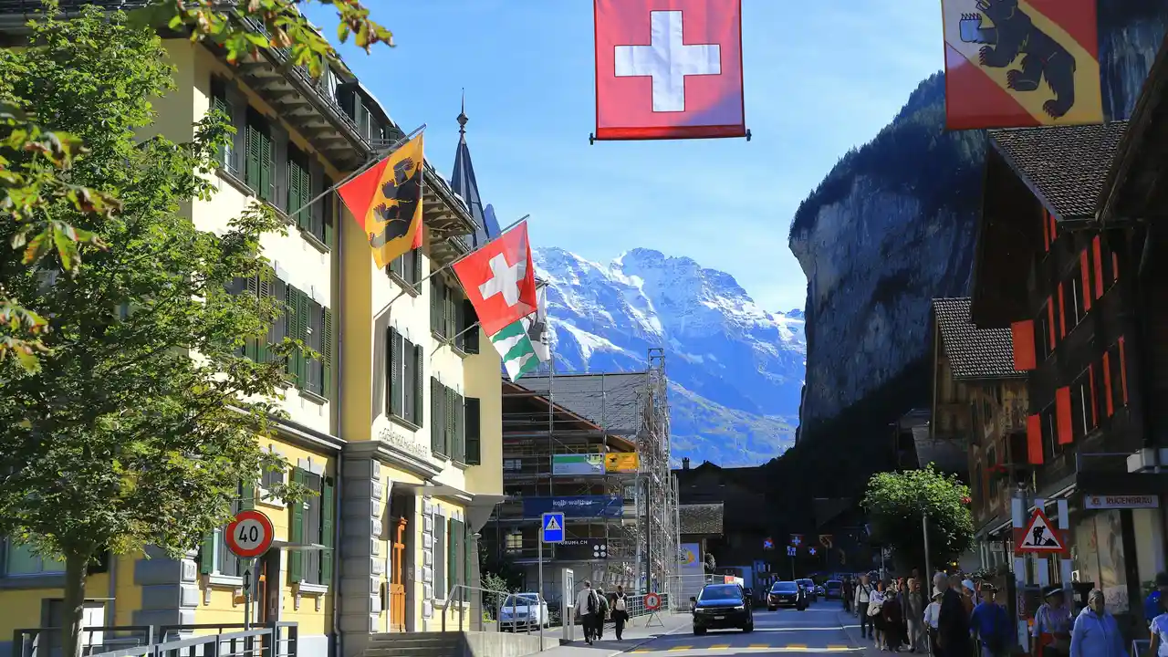 A vibrant street scene in a Swiss village, with traditional buildings adorned with the Swiss national flag and various cantonal flags featuring bear motifs. In the background, majestic snow-capped mountains rise sharply against a clear blue sky