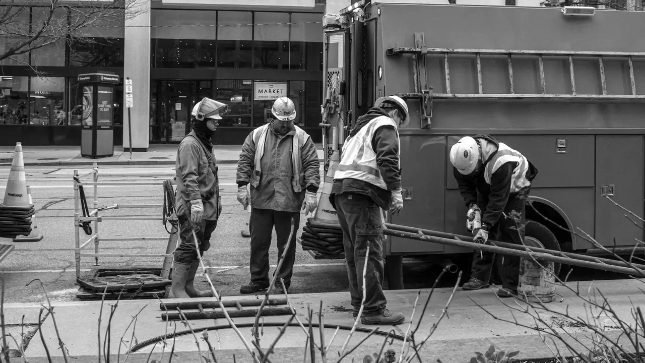A black-and-white photograph of four utility workers in hard hats and high-visibility vests working on a city sidewalk next to a service truck. One worker uses a tool on a piece of equipment while the others observe or assist, with a street and storefronts visible in the background.