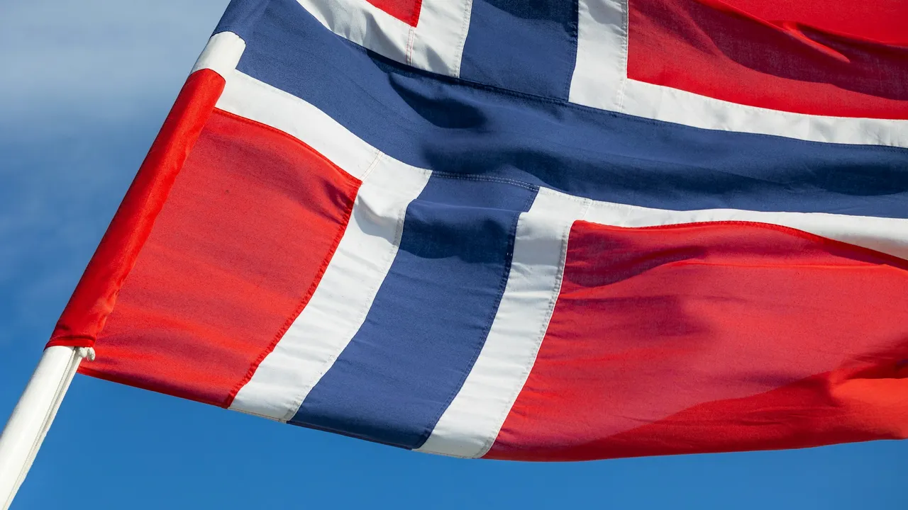 A close-up shot of the national flag of Norway, featuring a blue cross with a white outline on a red background, waving in the wind against a clear blue sky. The flag is brightly lit, showcasing its vibrant colors and texture.