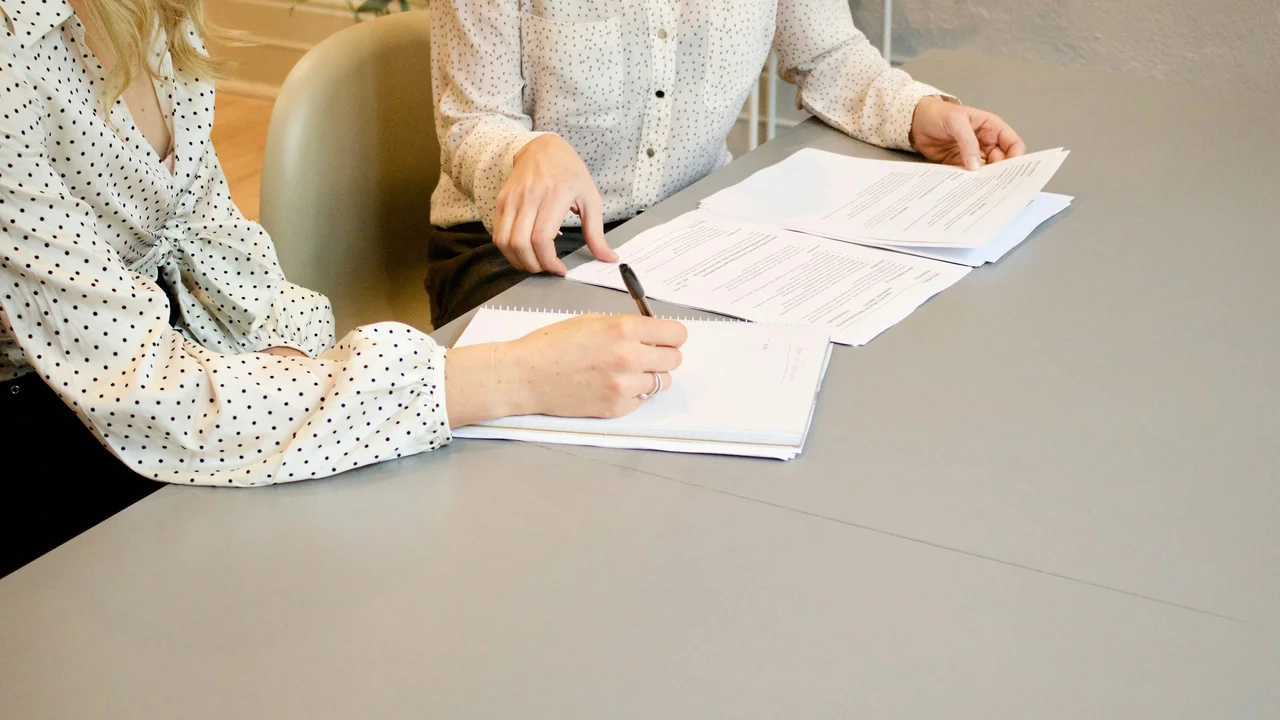 A professional close-up shot of a person’s hands neatly organizing a stack of official-looking documents on a wooden desk. A white graphic overlay on the left features the