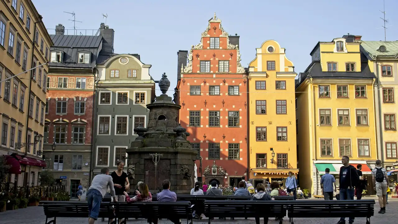 “Colorful historic buildings at Stortorget Square in Gamla Stan, Stockholm, Sweden, featuring traditional Nordic architecture, a central stone fountain, and people relaxing in a sunny old town plaza.