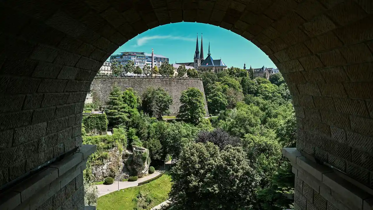 Scenic view of Luxembourg City old town and historic fortifications framed by a stone arch, overlooking lush green valleys and medieval architecture under a clear blue sky.