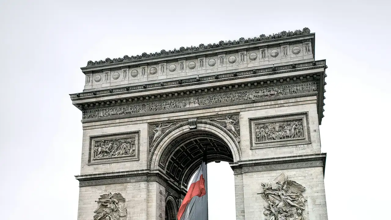 Arc de Triomphe monument in Paris, France, featuring detailed stone carvings and sculptures with the French flag hanging beneath the arch, symbolizing French history, national pride, and a popular tourist landmark.