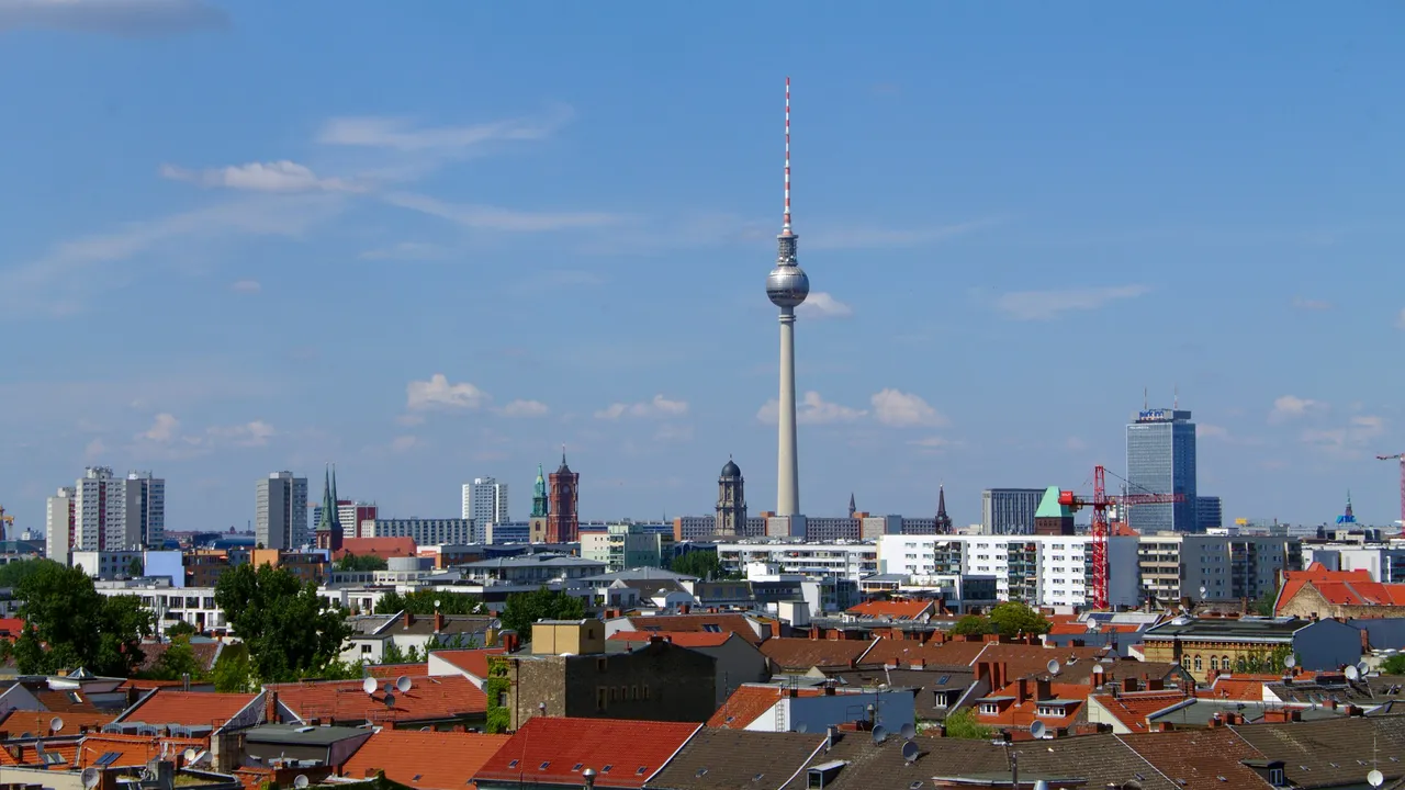 A wide view of the Berlin skyline under a clear blue sky, featuring the iconic Fernsehturm (TV Tower) rising above red-roofed buildings and urban blocks.