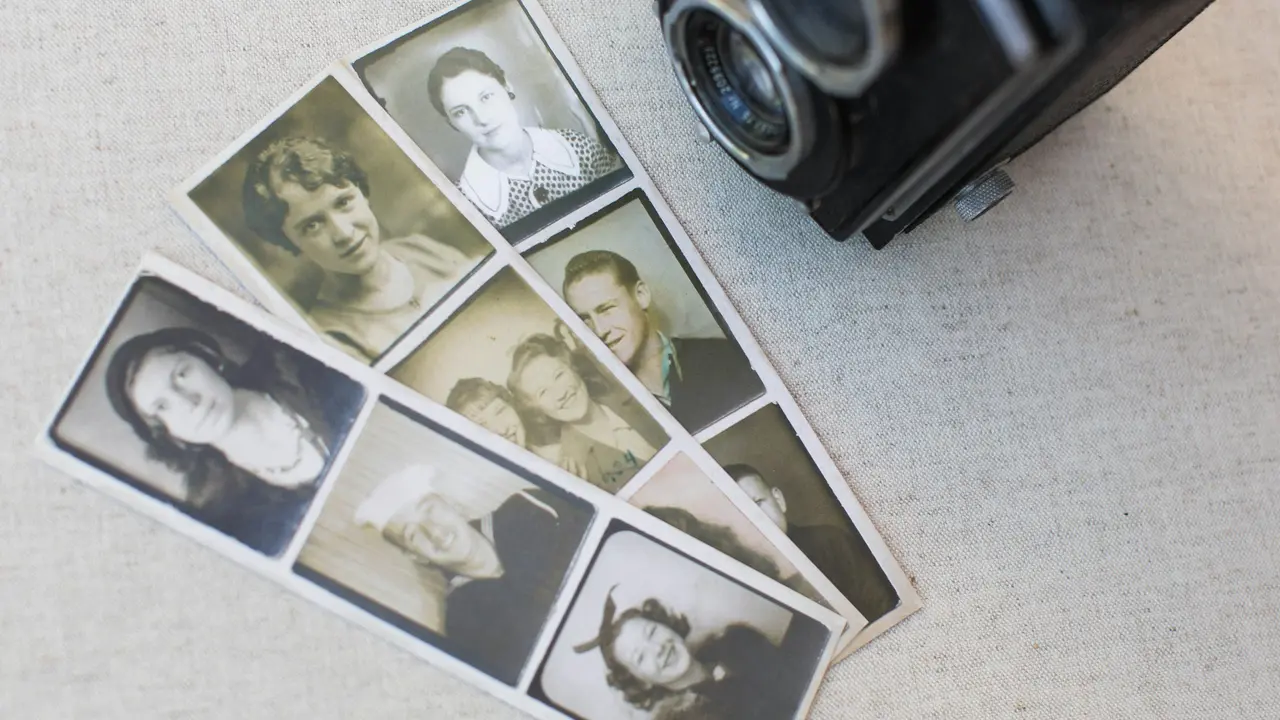  An image showing several strips of vintage black-and-white passport-style photographs laid out on a light-colored fabric surface. The small portraits feature various men and women in classic attire. A portion of a vintage twin-lens reflex (TLR) camera is visible in the upper right corner, resting near the photo strips.