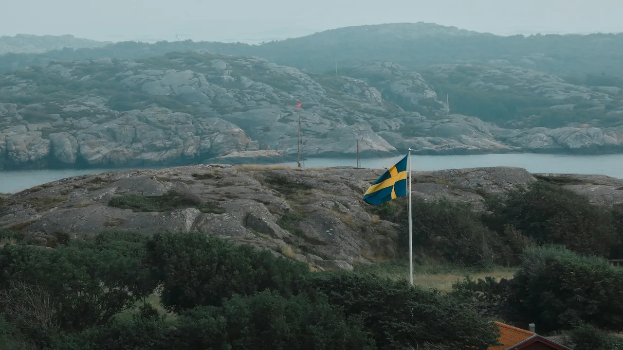 A Swedish flag flutters from a white flagpole in the foreground, set against a rugged coastal landscape of grey rocky hills and sparse green vegetation under a hazy sky