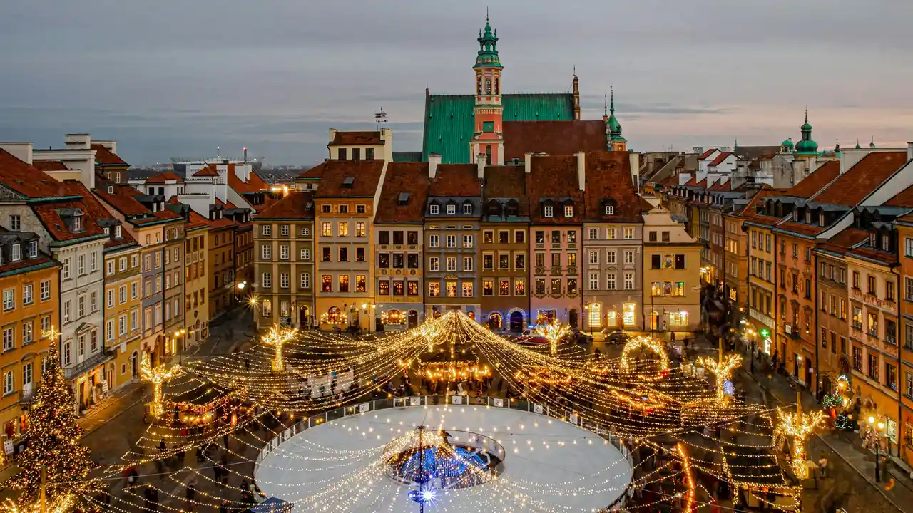 An aerial nighttime view of the circular Old Town Market Square in Warsaw, Poland, during the festive season. At the center of the square, a brightly lit ice skating rink surrounds the Warsaw Mermaid statue, which is illuminated by a canopy of overhead twinkling lights. The rink is bordered by colorful, historic tenement houses with gabled roofs, and several small wooden Christmas market stalls are visible along the perimeter.