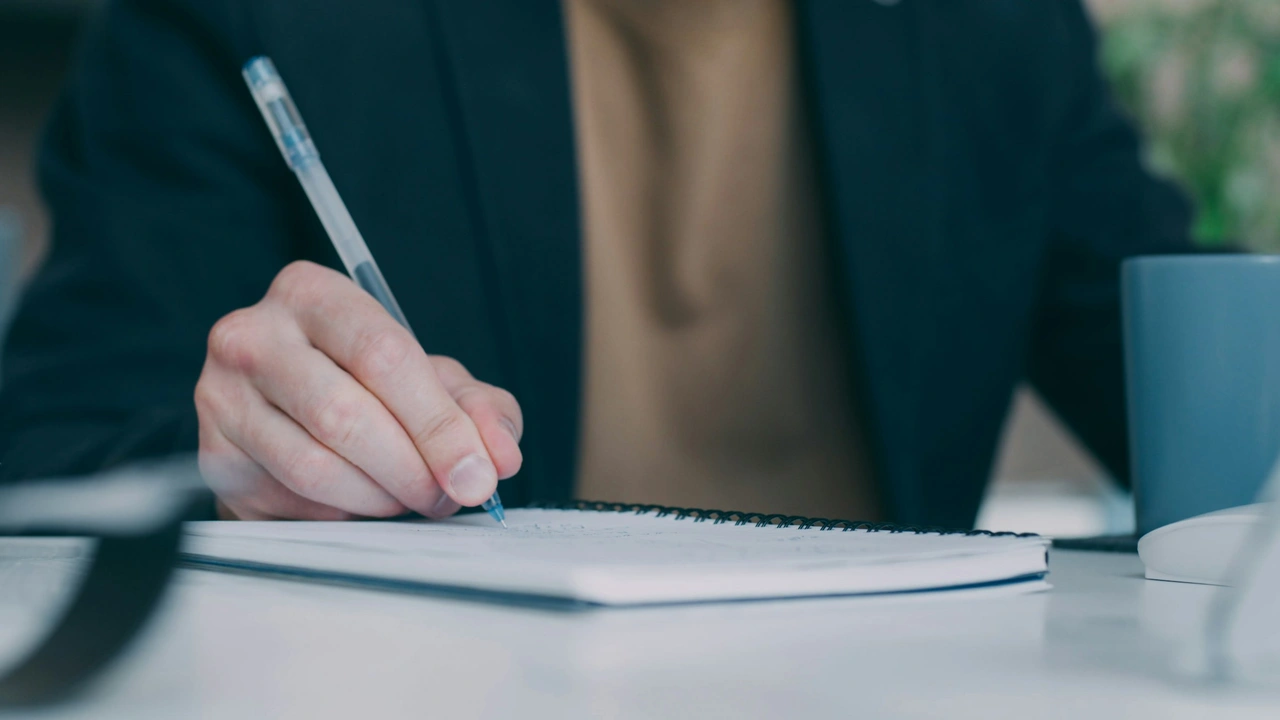 An image showing a person's hand holding a blue pen, poised to write in a white spiral-bound notebook. The person is wearing a dark blazer over a tan shirt, and the background is softly blurred, showing a desk with a grey mug.