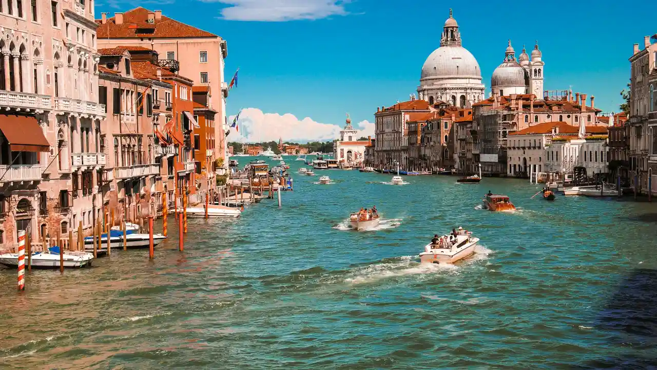 A vibrant, wide-angle shot of the Grand Canal in Venice, Italy, bustling with several motorboats and water taxis cruising through the turquoise water. The canal is lined with historic, multi-story buildings in warm shades of terracotta and cream. In the background, the iconic white domes of the Santa Maria della Salute basilica stand prominently against a bright, clear blue sky.