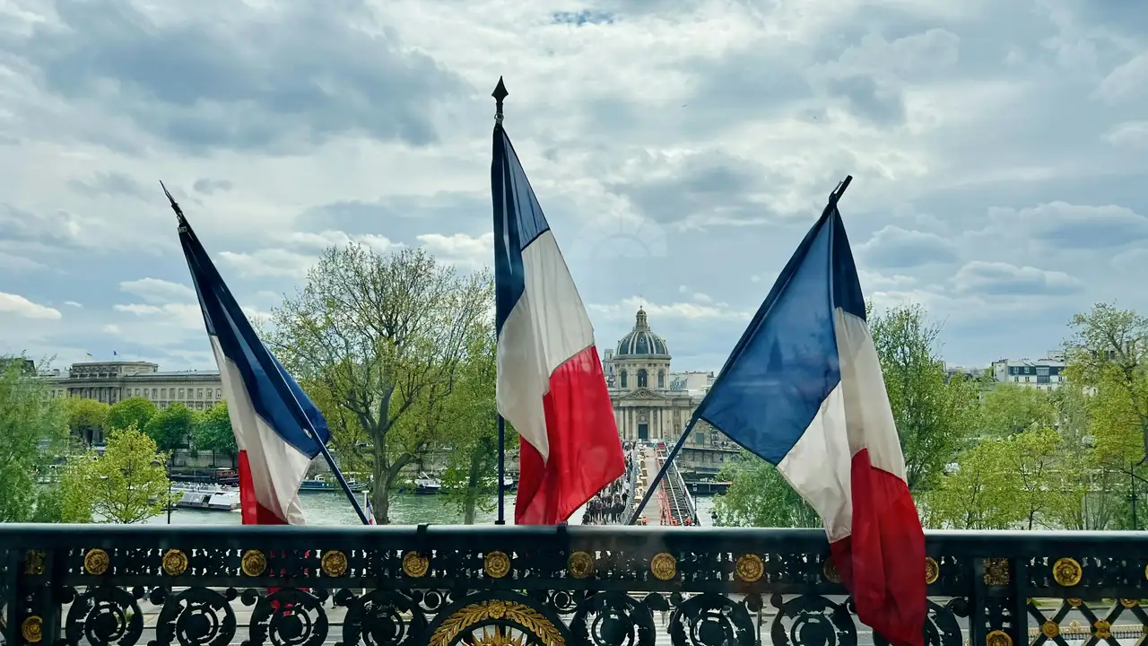 A view from a balcony featuring three French national flags (blue, white, and red tricolor) blowing in the wind. In the background, a pedestrian bridge crosses the Seine River, leading toward the historic Institut de France building with its prominent dome under a cloudy Parisian sky. The balcony features an ornate black and gold wrought-iron railing.