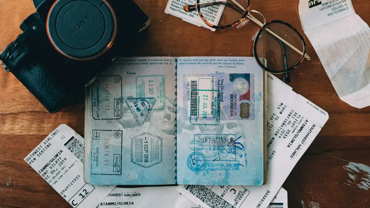 An overhead shot of a wooden table scattered with travel essentials. In the center is an open passport filled with colorful entry and exit stamps and an Indonesian visa. Surrounding it are several Southwest Airlines boarding passes, a black Sony camera, a pair of wire-rimmed glasses, and paper receipts.