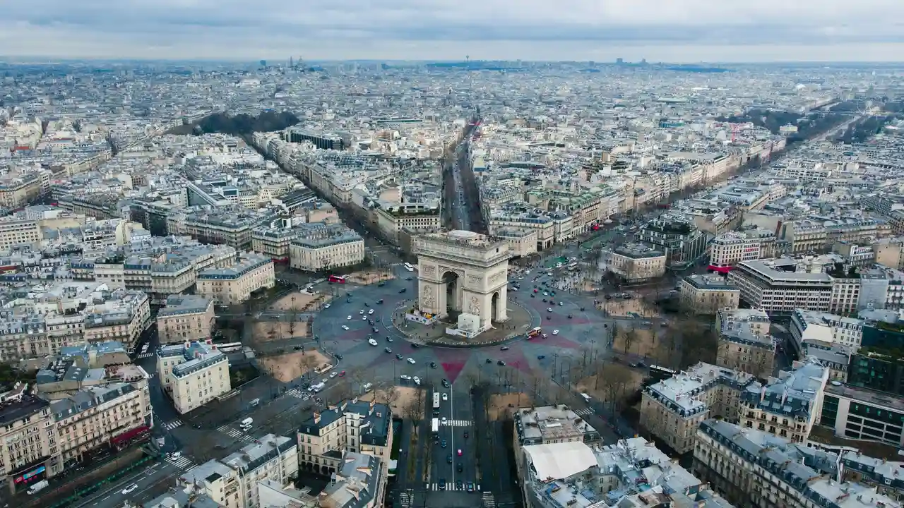 An aerial, high-angle view of the Arc de Triomphe in Paris, situated at the center of the Place Charles de Gaulle. Multiple roads radiate outward from the monument into the dense, historic cityscape of Paris under a soft, overcast sky. Traffic is visible circling the monument and moving along the wide avenues
