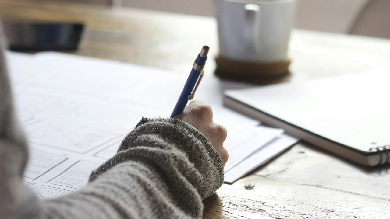 A person filling out a visa application form by hand at a desk, holding a pen over official documents with a notebook and coffee cup nearby, representing the visa application process.