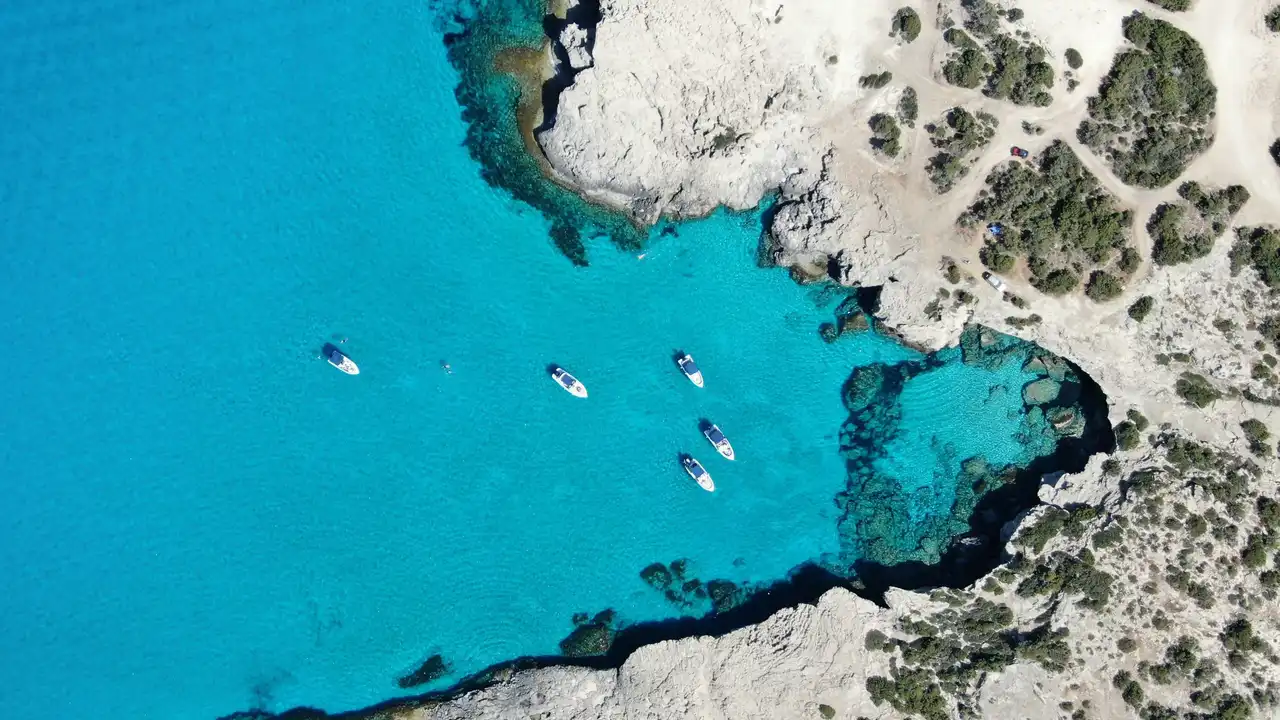 Aerial view of a turquoise coastline in Cyprus featuring crystal-clear Mediterranean waters, rocky cliffs, small boats anchored in a scenic bay, and rugged natural shoreline.