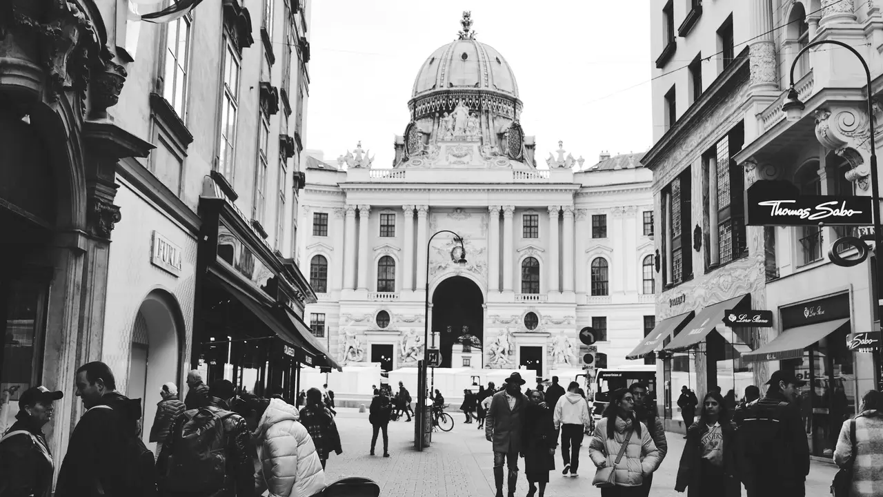 Black and white street scene in Vienna, Austria, showing pedestrians walking toward the historic Hofburg Palace with its domed architecture, surrounded by narrow shopping streets and classic European buildings.