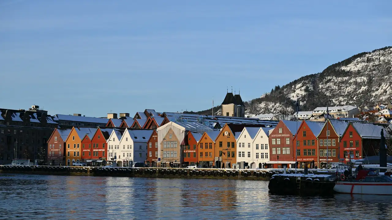  A row of colorful, snow-capped historic wooden buildings along a waterfront in Bergen, Norway, with a snowy mountain in the background
