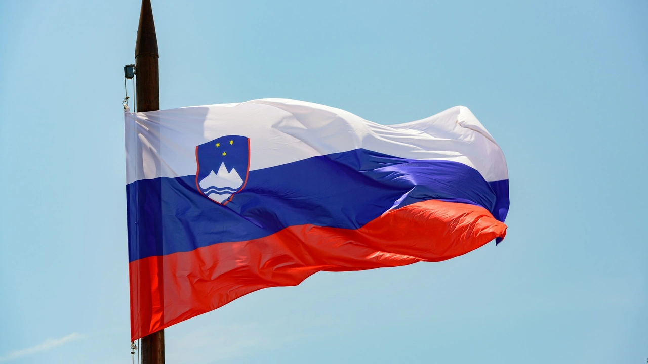 A medium shot of the national flag of Slovenia billowing in the wind against a clear, pale blue sky. The flag features three equal horizontal bands of white, blue, and red, with the Slovenian coat of arms positioned in the upper left corner.