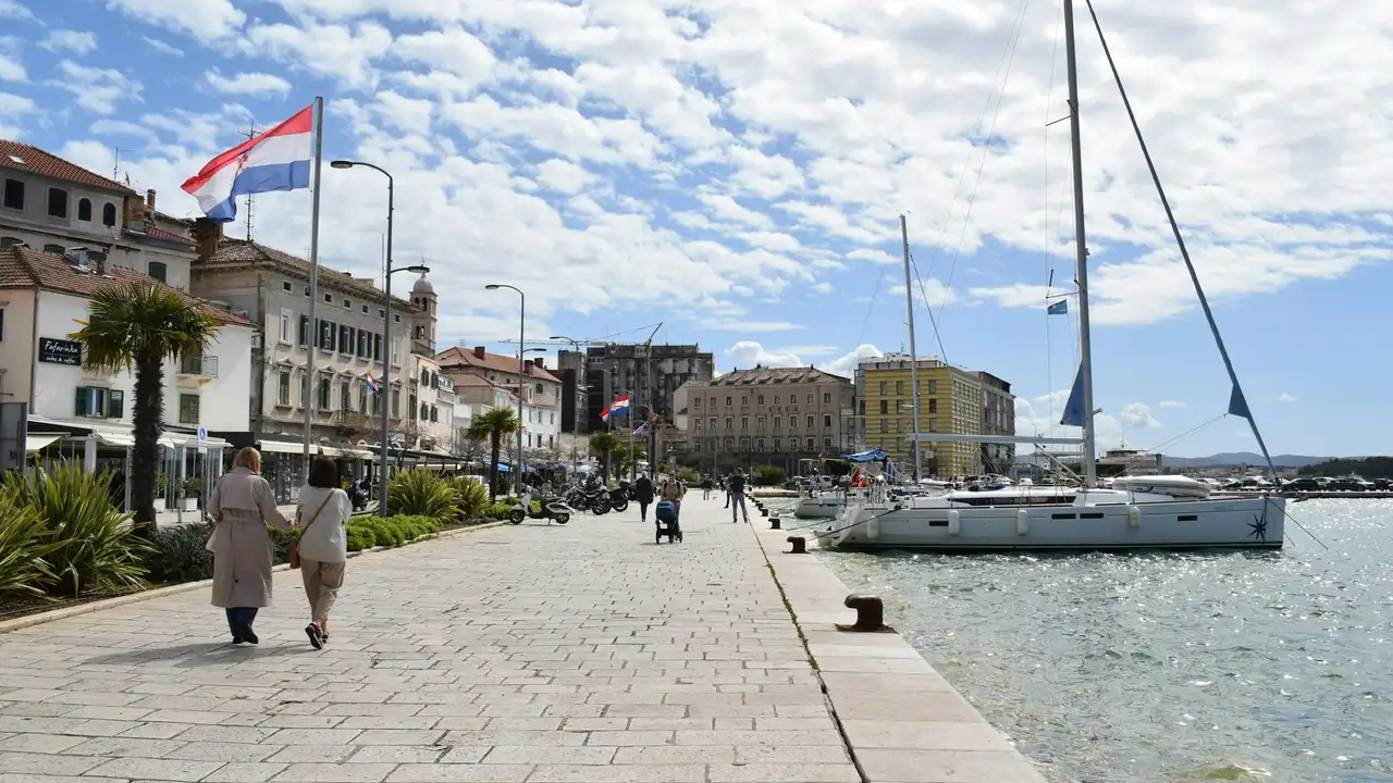 A wide stone promenade in a coastal Croatian town under a bright, cloudy sky. To the left, two people walk past historic multi-story buildings, palm trees, and cafes flying the Croatian flag. To the right, several sailboats are moored in the sparkling blue water of the harbor