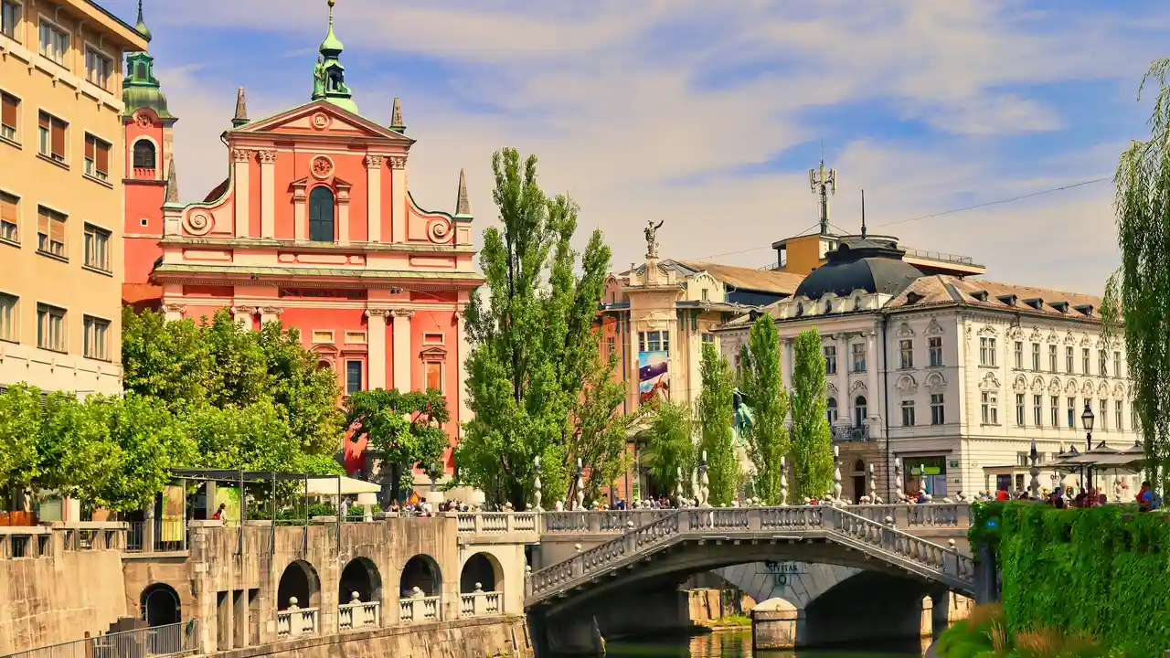 A vibrant, wide-angle shot of the Triple Bridge crossing the Ljubljanica River in Ljubljana, Slovenia. In the background, the salmon-pink facade of the Franciscan Church of the Annunciation stands prominently against a bright blue sky with wispy clouds. The scene is lined with lush green trees and historic European architecture, with stone balustrades and pedestrian walkways overlooking the calm water.