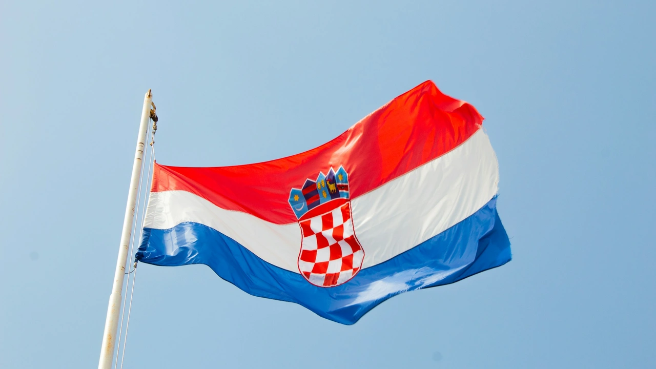 The national flag of Croatia waving against a clear, light blue sky. The flag features three horizontal stripes of red, white, and blue, with the Croatian coat of arms—a red and white checkered shield topped by a crown of five smaller shields—centered on the white stripe.