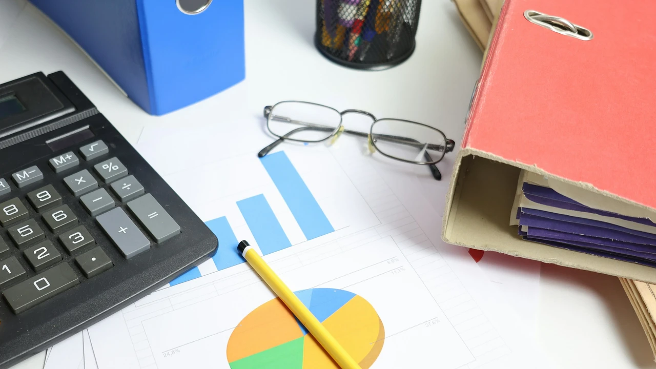 An office desk featuring a calculator, a yellow pencil resting on financial charts (bar and pie graphs), a pair of eyeglasses, and several large file binders in blue and red