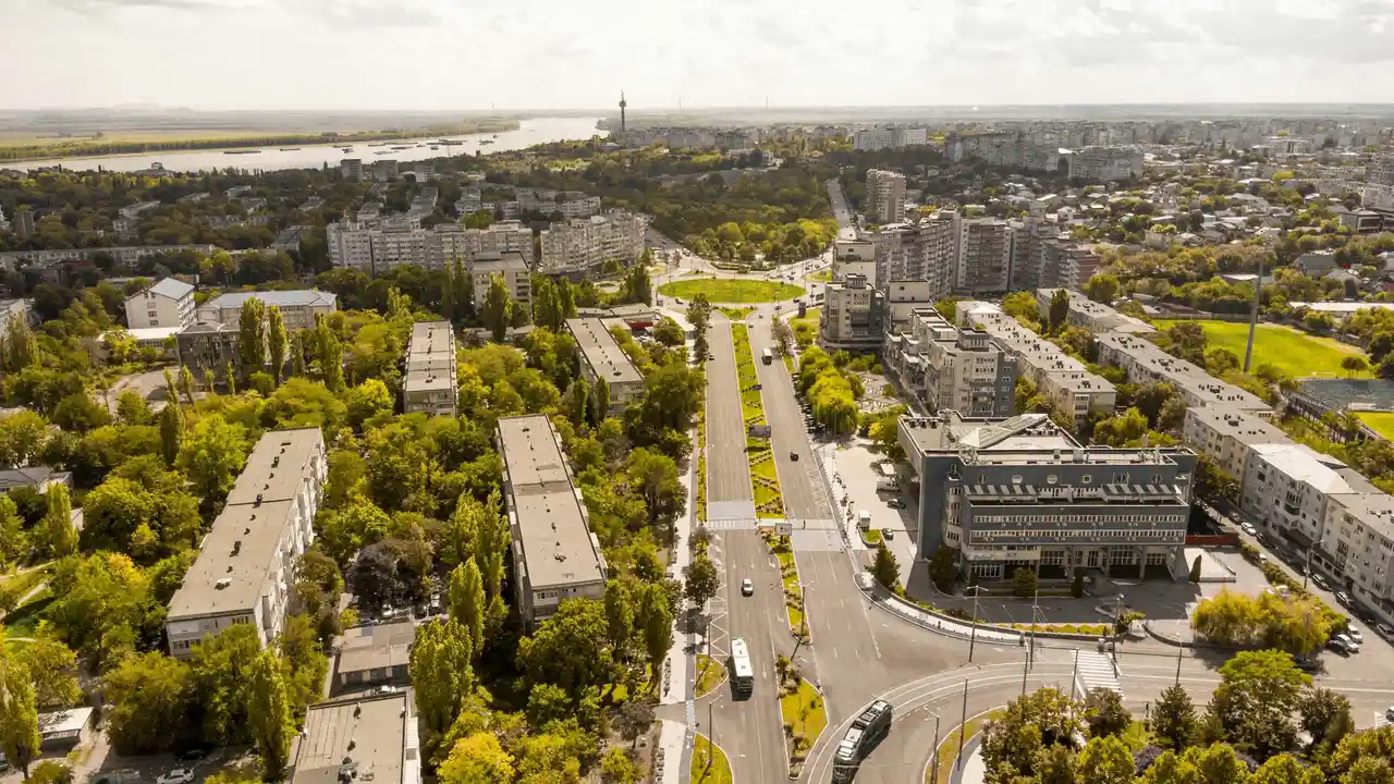 An aerial, high-angle view of a sunlit city in Romania, featuring a wide boulevard with a green median that leads toward a large roundabout. Lush green trees are interspersed among several multi-story apartment buildings and commercial structures. In the background, a wide river flows along the edge of the city under a hazy, bright sky.