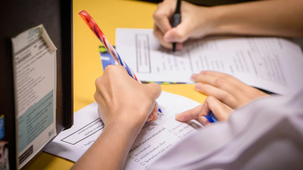 A close-up, over-the-shoulder shot shows two people sitting at a bright yellow table, focused on filling out official forms. In the foreground, one person holds a red and white striped pen, poised to write on a document featuring checkboxes and lines of text.