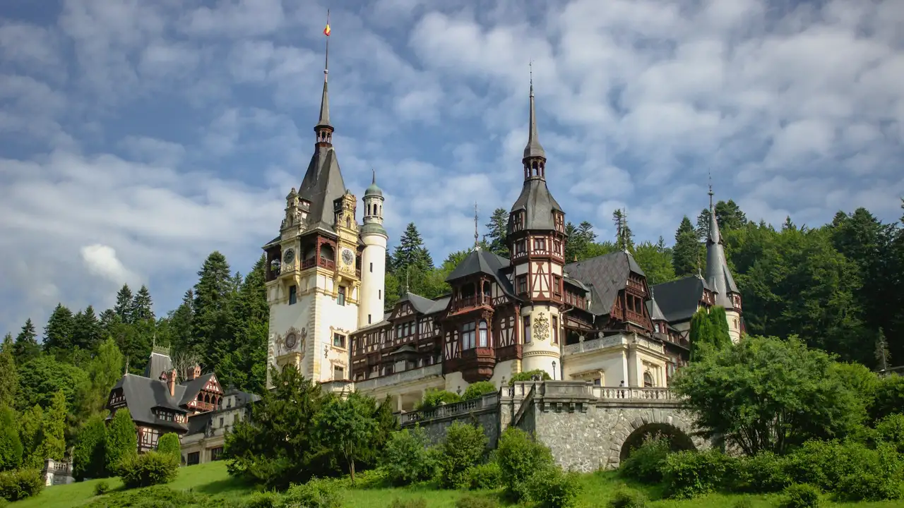 A wide shot of Peleș Castle in Romania, showcasing its ornate Neo-Renaissance architecture with tall spires, timber-framed walls, and intricate balconies. The castle is nestled on a lush green hillside surrounded by dense evergreen trees under a bright blue sky with soft white clouds.