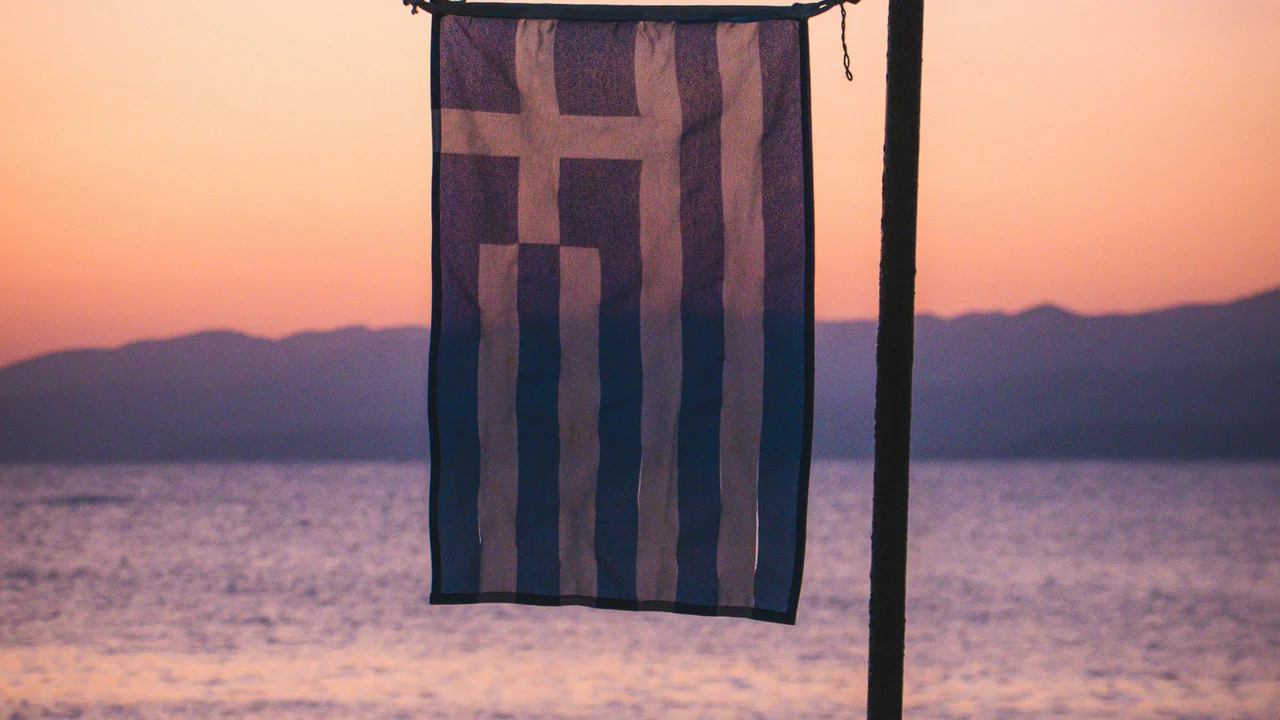 A vertical Greek flag hangs from a pole in the foreground, partially silhouetted against a soft sunset. The background features the calm waters of the sea and the dark, layered outlines of distant mountains under a sky glowing with warm orange and pink hues