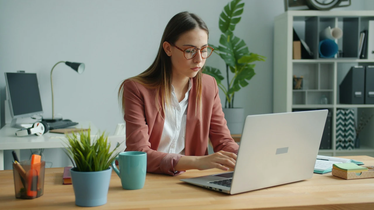 A young woman wearing glasses and a pink blazer sits at a wooden desk, focused on her laptop. Her workspace is organized with a teal mug, a small potted plant, and a white bookshelf in the background.