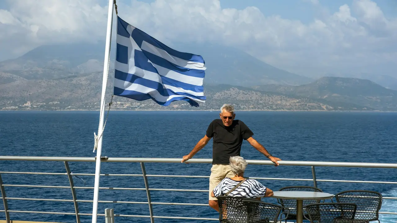 A large Greek flag waves in the wind on the deck of a boat. An older man in a black t-shirt leans against the railing next to a seated woman, with the blue sea and hazy mountains visible in the distance.
