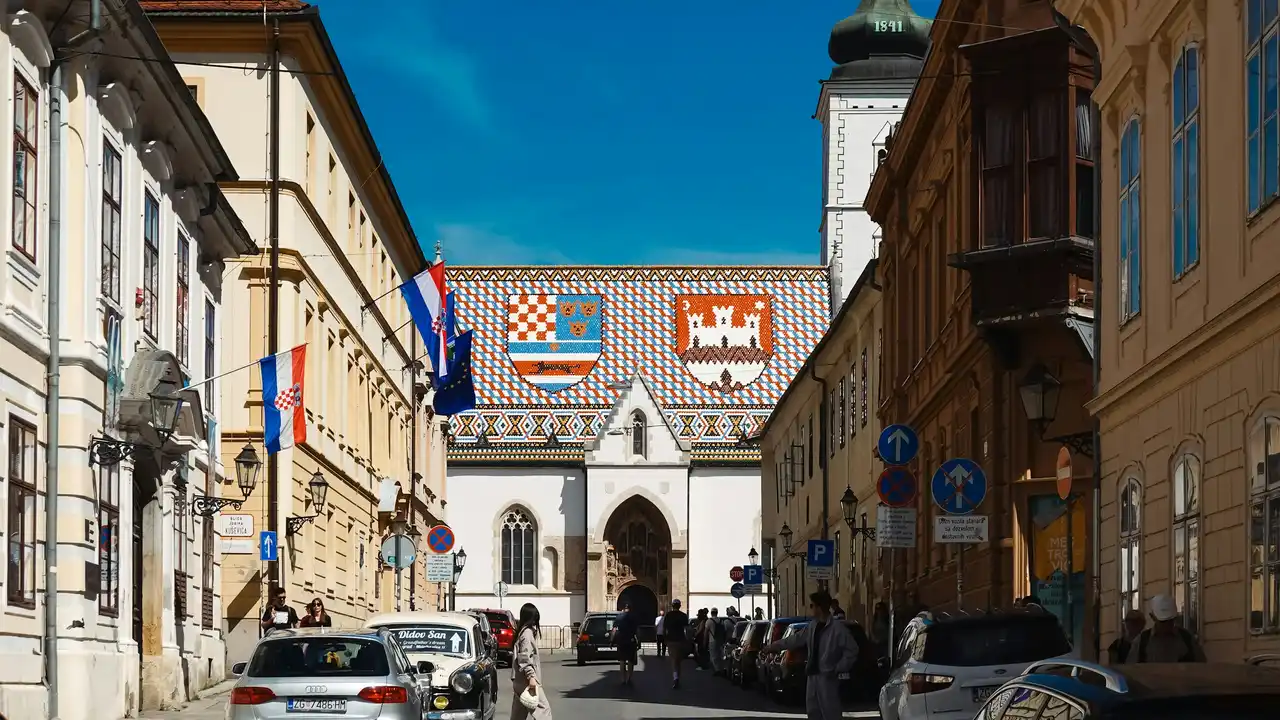 A vibrant view of St. Mark's Church in Zagreb, Croatia, framed by historic city buildings and parked cars. The church is famous for its colorful tiled roof featuring the medieval coat of arms of Croatia, Dalmatia, and Slavonia on the left, and the emblem of Zagreb on the right. Several Croatian flags hang from a building on the left under a clear blue sky.