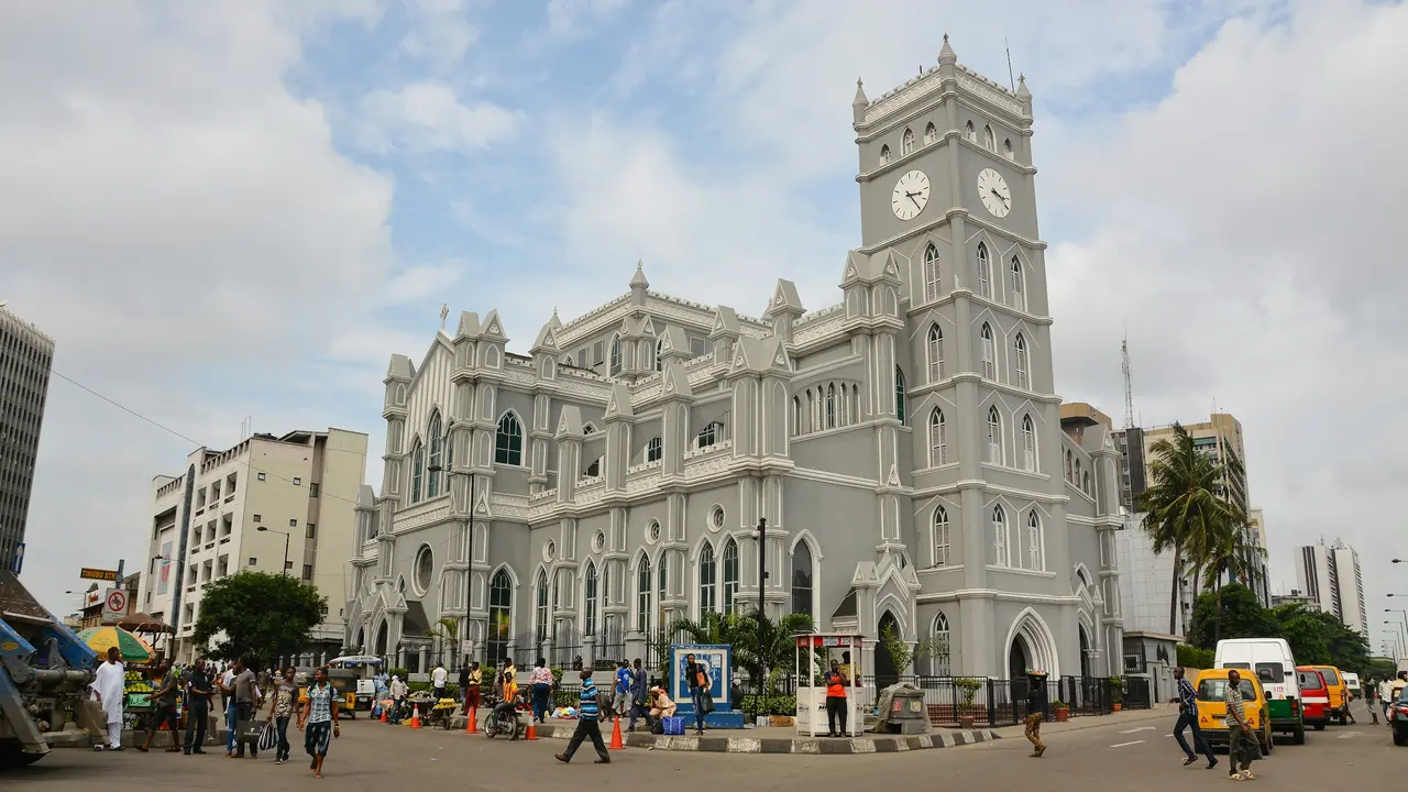 Holy Cross Cathedral in Lagos, Nigeria, a historic grey Gothic-style church with a prominent clock tower, surrounded by busy city streets, pedestrians, and urban traffic under a partly cloudy sky.