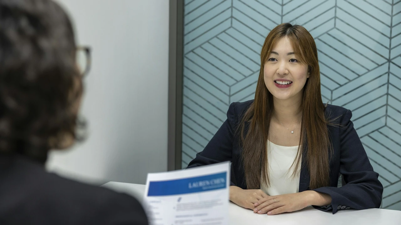 A woman in business attire smiles while sitting at a desk during a visa or job interview, facing an interviewer who is holding documents in a modern office setting.