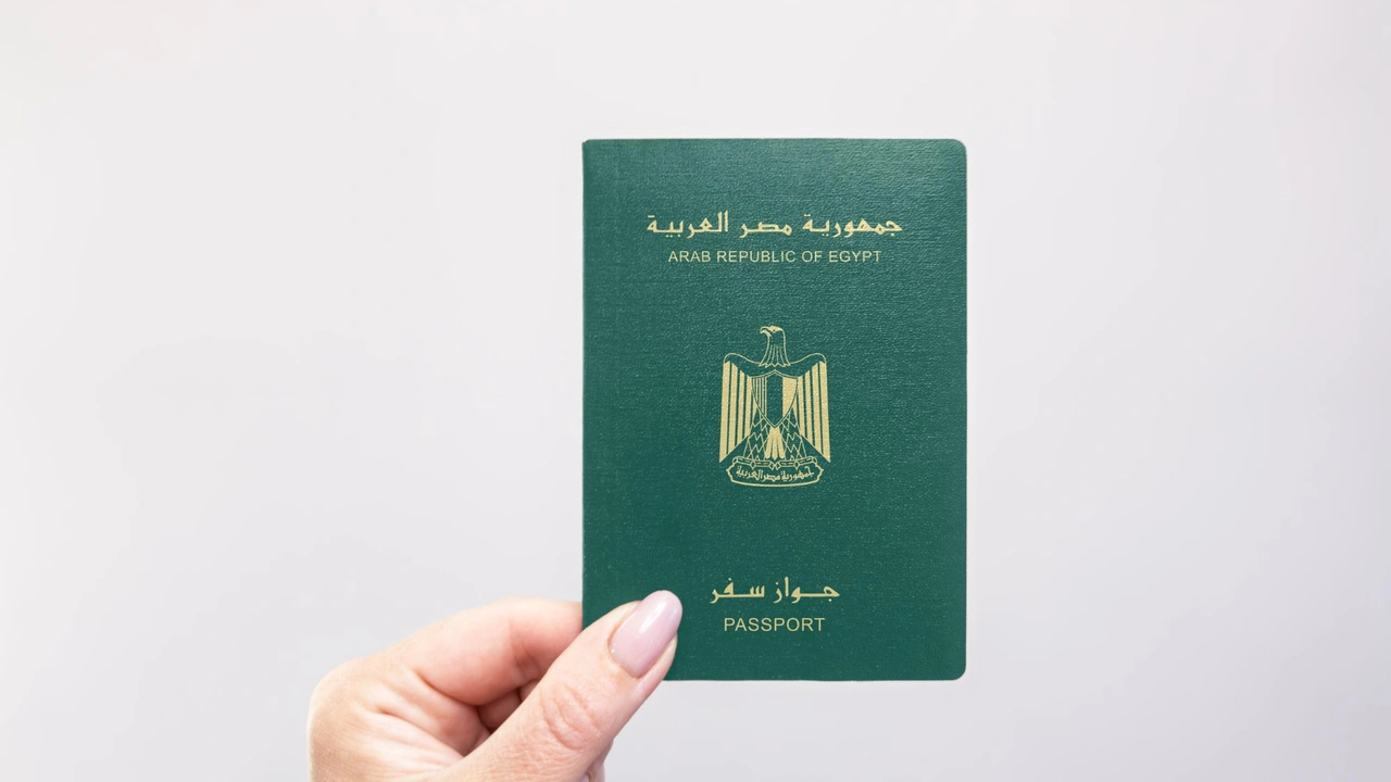 Hand holding an Egyptian passport with gold emblem and “Arab Republic of Egypt” text on a green cover against a neutral background.