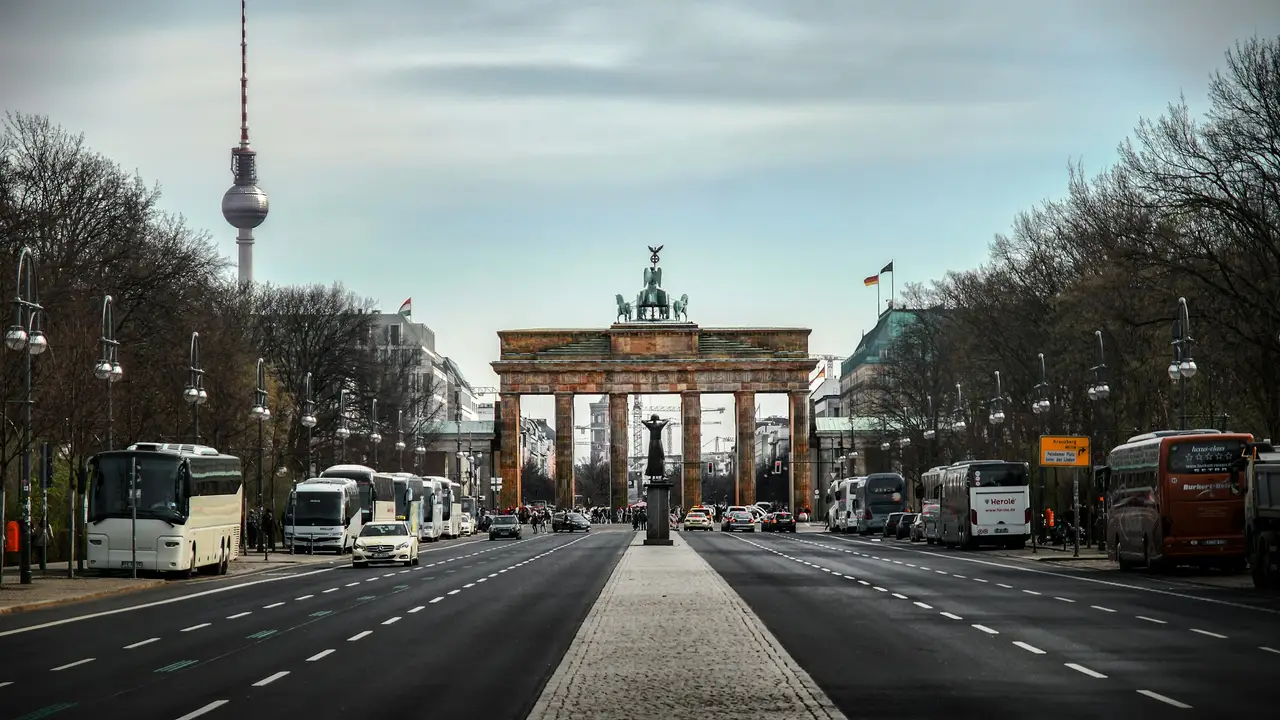 A wide-angle, symmetrical shot of the Brandenburg Gate in Berlin, Germany, centered at the end of a wide, multi-lane road. The neoclassical monument is topped by the Quadriga statue, and to its left, the Berlin TV Tower (Fernsehturm) rises into a pale, overcast sky.