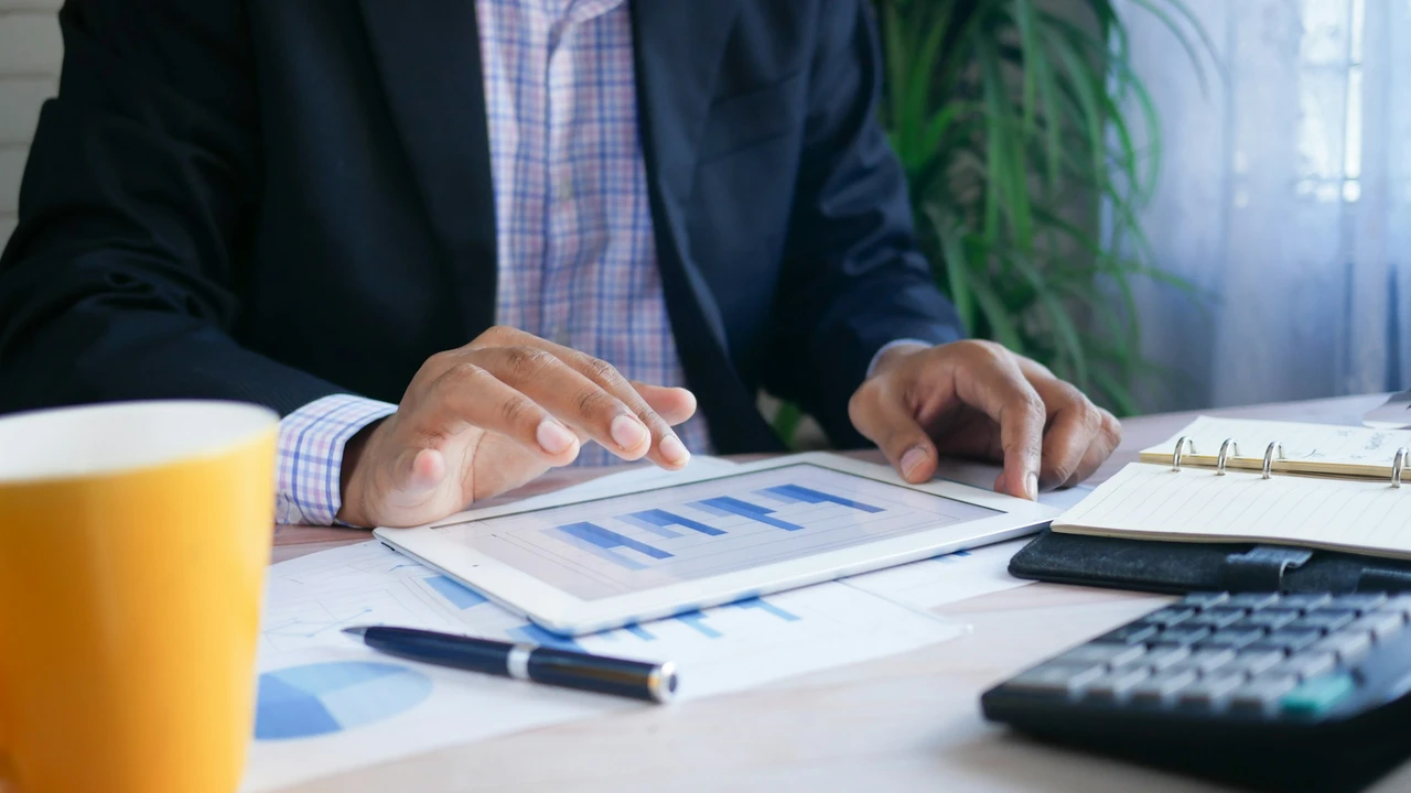A close-up, high-angle shot of a person’s hands using a white tablet to review vibrant financial charts and graphs. On a wooden desk surrounding the tablet are various business essentials, including a calculator, a pair of glasses, a white coffee cup, and printed financial documents with additional data visualizations. The lighting is bright and professional, highlighting a focused, data-driven workspace.