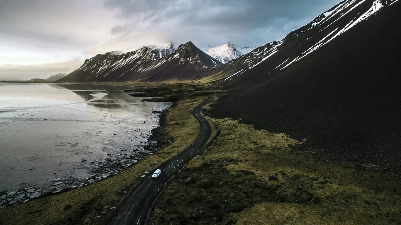 An aerial, wide-angle shot of a white vehicle driving along a winding coastal road in Iceland. The road curves between a dark, rugged mountain slope on the right and a partially frozen lagoon with floating ice on the left. In the distance, dramatic snow-capped mountain peaks rise under a moody, overcast sky, capturing the vast and wild nature of the Icelandic landscape